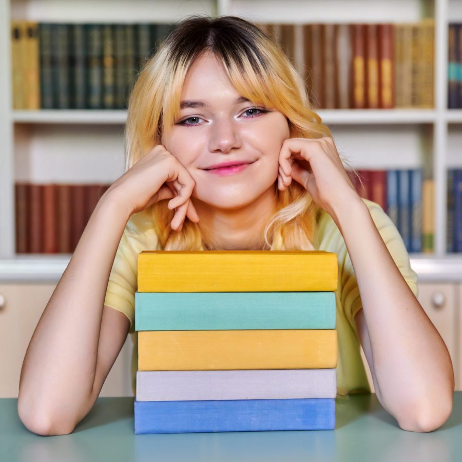 Smiling pretty female teenage high school student looking at camera with stack of books in library. Back to school, back to college, knowledge, learning, education, teens concept